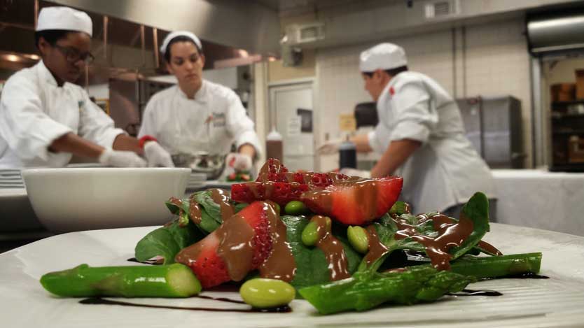 A strawberry spinach salad sits in the foreground as culinary students work in the background.