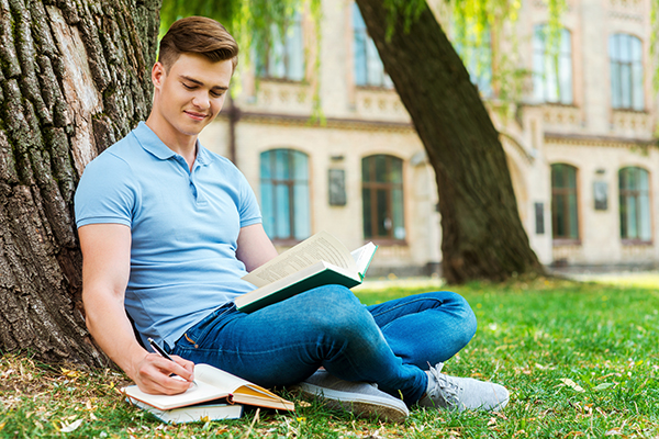 white male sitting next to a tree on a college campus studying