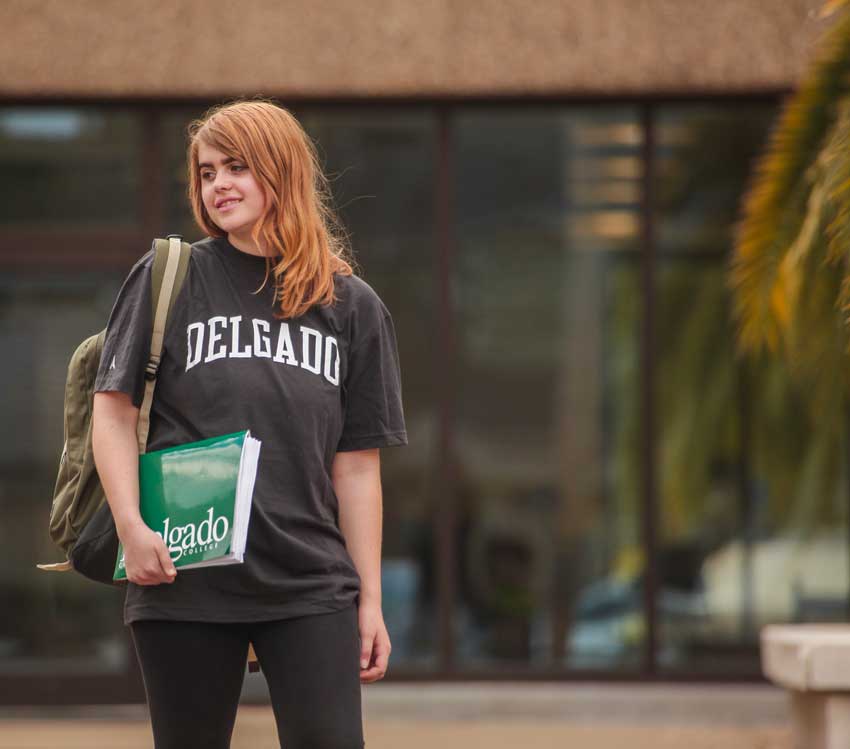 A young female Delgado student wearing a Delgado t-shirt poses in front of an academic building holding a Delgado branded folder and wearing a backpack.